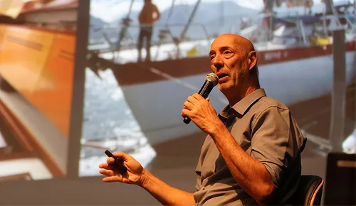 Beto Pandiani ao microfone em um workshop, com a imagem de um veleiro projetada atrás